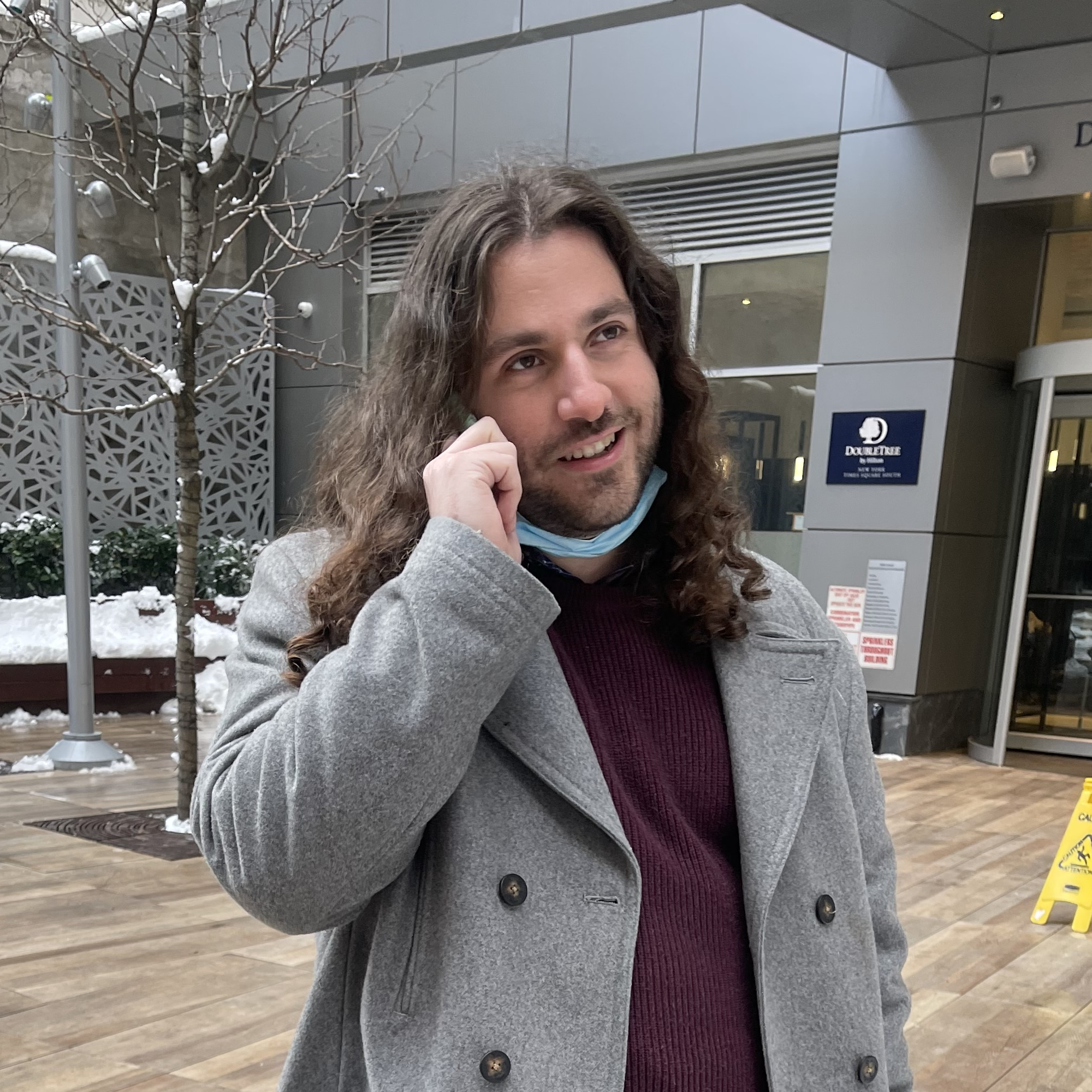 A picture of me, CJ Axisa, standing in New York City wearing a gray jacket with a maroon sweater underneath while talking on the phone and smiling at a 3/4 view.